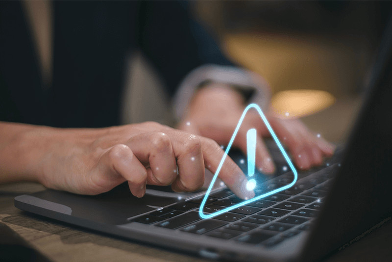 person typing on a laptop with a blue warning sign over it